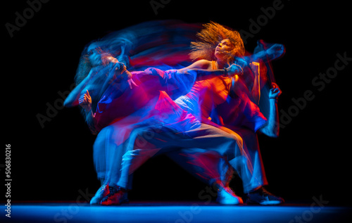Young man and woman dancing hip-hop in sportive style clothes on dark background at dance hall in mixed neon light. Youth culture, hip-hop, movement