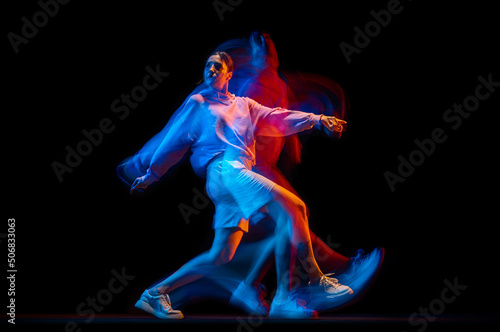 Young woman dancing hip-hop in sportive style clothes isolated on dark background at dance hall in mixed neon light. Youth culture, hip-hop, movement