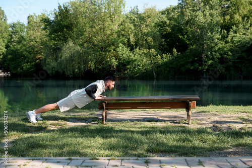 Young brunette man do morning exercises in city park pushing his body up from parks' bench