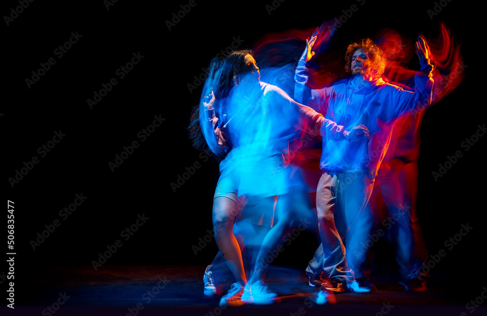Fototapeta premium Young man and woman dancing hip-hop in sportive style clothes on dark background at dance hall in mixed neon light. Youth culture, hip-hop, movement