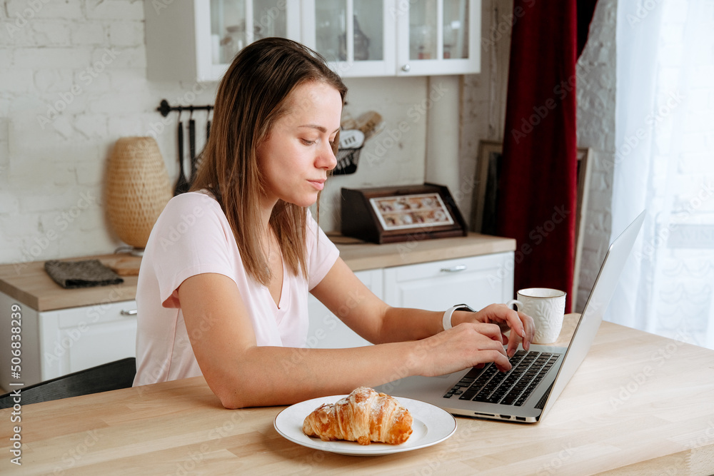 Young woman eating breakfast at home and working on her laptop in her kitchen, looking at the screen Young woman eating breakfast at home and working on her laptop in her kitchen, looking at the screen