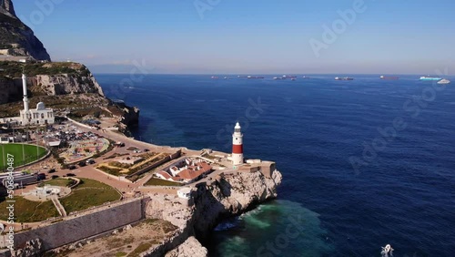 Red and white striped lighthouse and a large mosque at Europa point in Gibraltar overlooking several freighters at anchor as the Mediterranean swell splashes against the cliffs. Drone panning shot