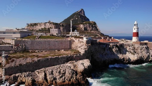 Waves from the clear blue Mediterranean splash against the cliffs below the recognizable lighthouse at Europa Point against the backdrop of Gibraltar's upper rock. Close up drone lowering shot