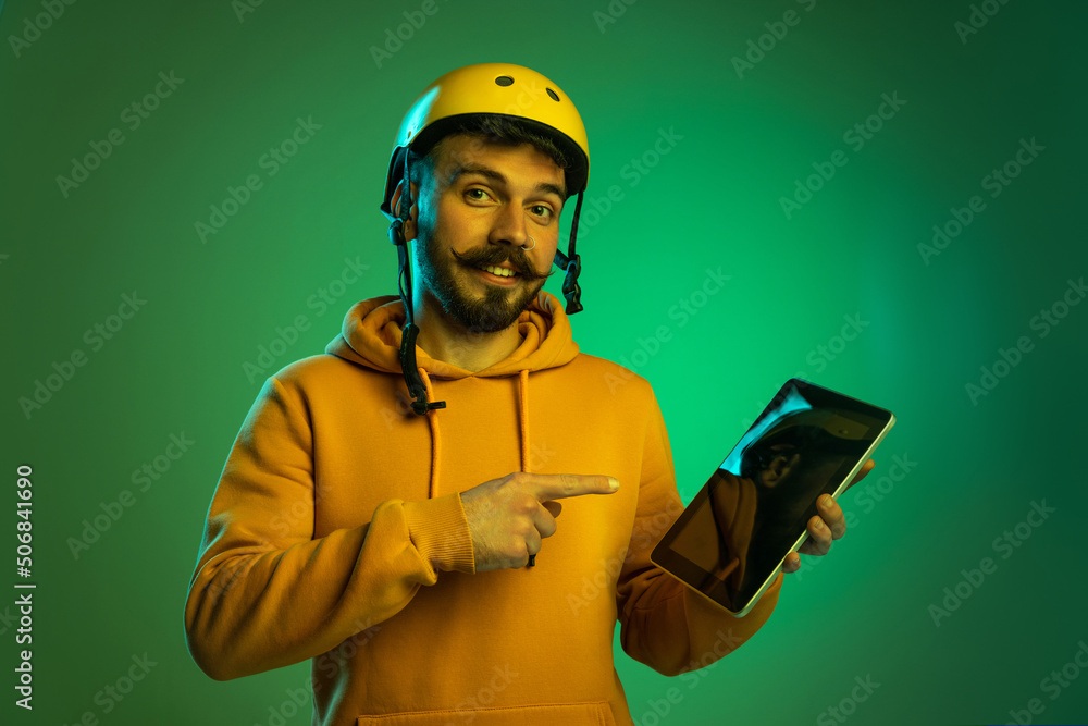 Portrait of young man working on delivery service, holding tablet isolated over green studio background in neon light. Devices dervice Portrait of young man working on delivery service, holding tablet isolated over green studio background in neon light. Devices dervice
