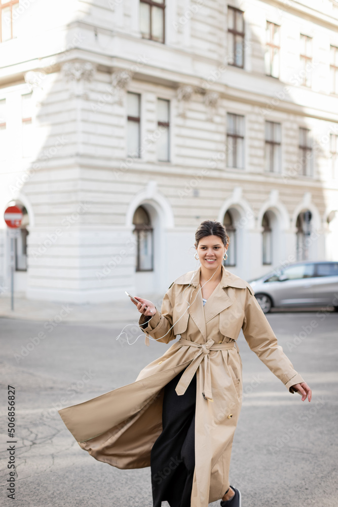 cheerful woman in wired earphones holding mobile phone and listening music on street in vienna.