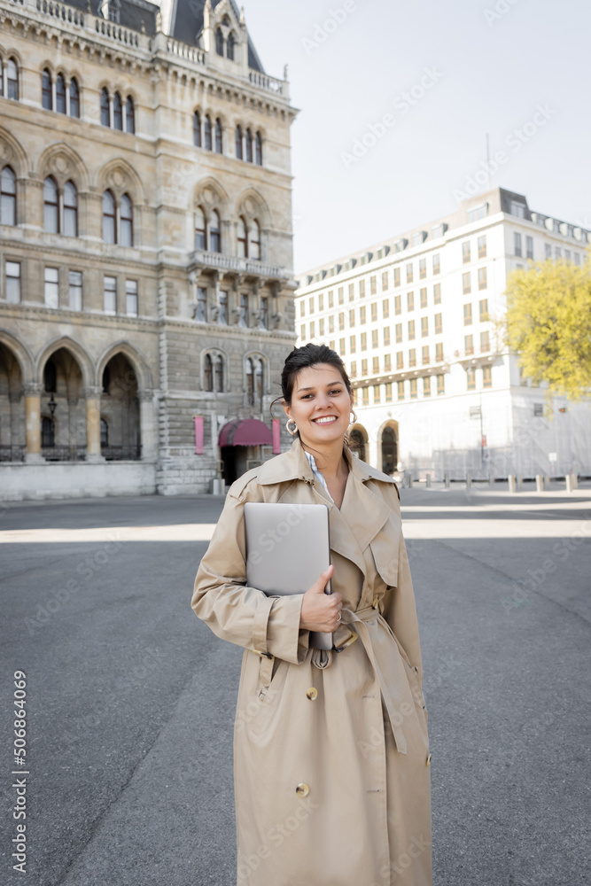 happy freelancer in beige trench coat walking with laptop on street of vienna.