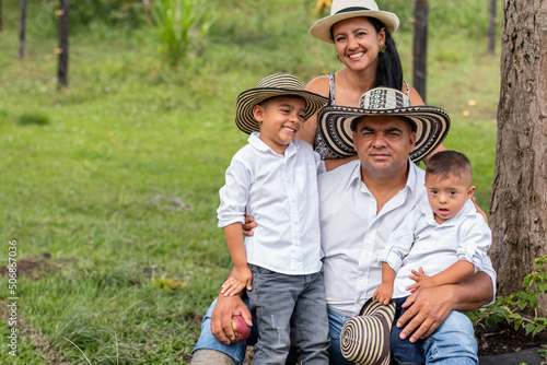 Portrait of a beautiful colombian family with brown skin. Dad, wife and children hug and look at the camera. People wearing hats in the countryside. Son with intellectual disability, trisomy.