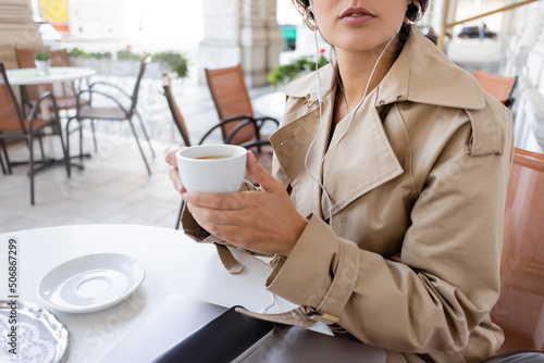 Photography cropped view of woman in trench coat listening music and holding cup near smartphone and laptop in cafe terrace