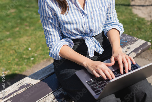 cropped view of woman typing on laptop keyboard while sitting on bench.