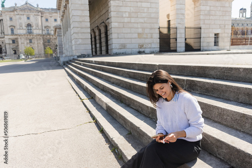 smiling woman listening music in wired earphones and using smartphone while sitting on stairs.