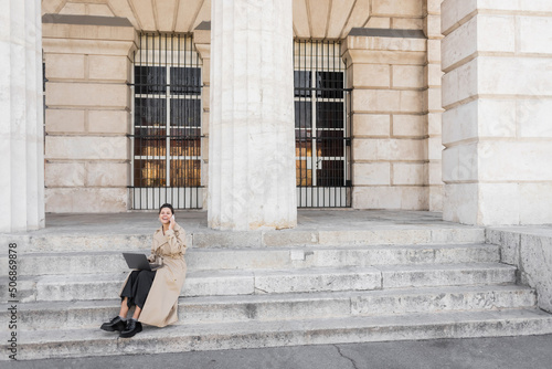 joyful woman in trench coat sitting on concrete stairs  talking on smartphone and working remotely on laptop in vienna.