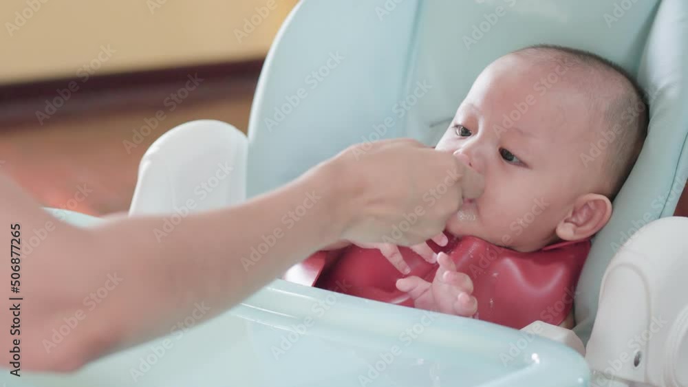 Authentic shot of cute asian newborn baby eating meal sitting on high chair. While hand of mother feeding healthy food give to child with spoon. Innocent little infant adorable. Mother Day concept. Authentic shot of cute asian newborn baby eating meal sitting on high chair. While hand of mother feeding healthy food give to child with spoon. Innocent little infant adorable. Mother Day concept.