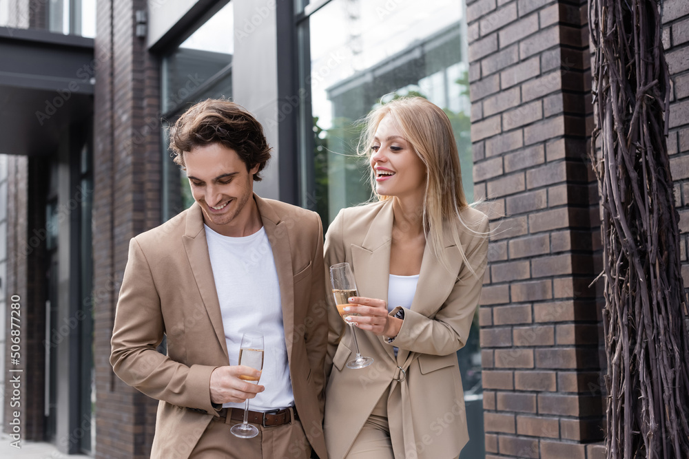 joyful young couple in beige suits walking with champagne glasses on city street.