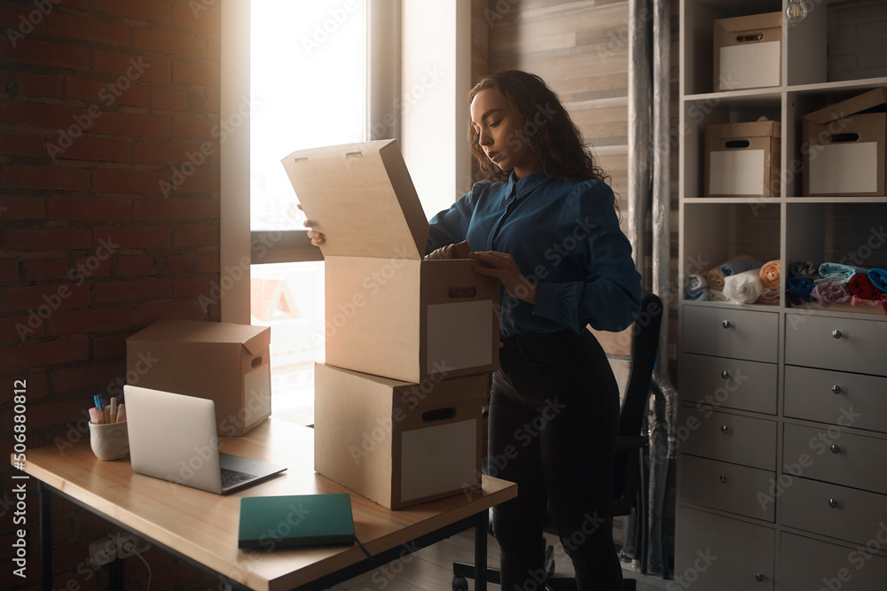 Fototapeta premium Young Asian curly-haired logistics worker packs goods, collects parcels, puts things in a carton box, prepares a parcel for shipment, transports, collects donations, business owner, delivery