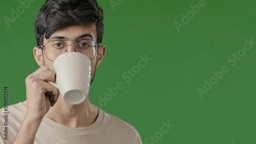 Close up portrait of arabian indian handsome young man millennial guy stand on green background drink coffee hot herbal tea cold water spend free time hispanic male daydreamer rest has morning routine photo