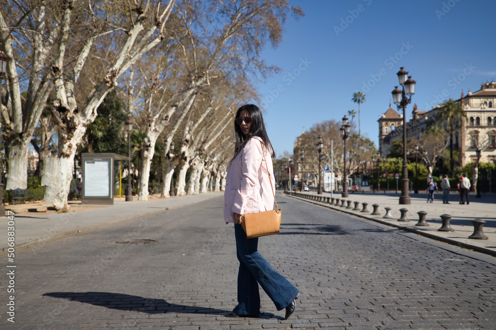 Young beautiful South American woman in a pink jacket, jeans, brown handbag and sunglasses, on vacation in Europe, crossing a city street. Concept vacation, travel, beauty, fashion.