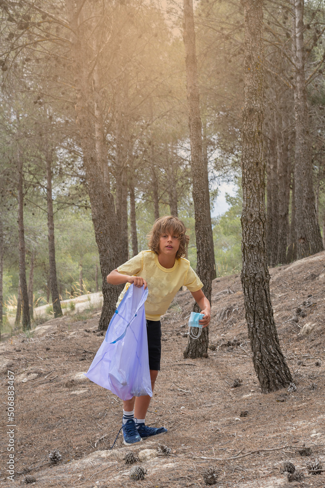 blond boy in yellow T-shirt collects rubbish and plastic in the forest, carrying a purple rubbish bag. blond boy in yellow T-shirt collects rubbish and plastic in the forest, carrying a purple rubbish bag.