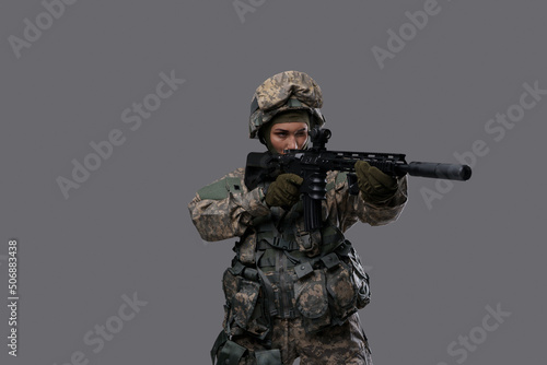 Portrait of militant woman dressed in camouflage clothes aiming rifle against gray background.