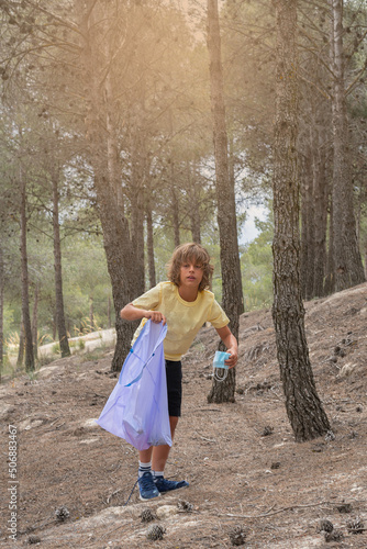 blond boy in yellow T-shirt collects rubbish and plastic in the forest, carrying a purple rubbish bag.