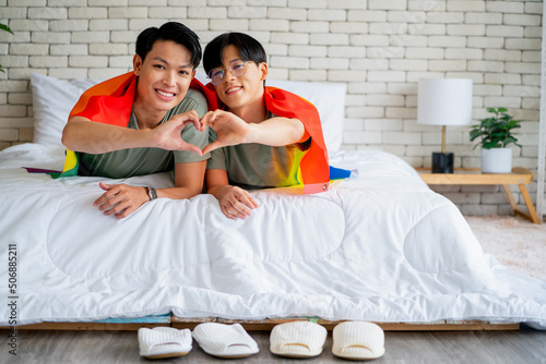 Couple gay or lgbt man with rainbow flag covering use hand to show heart symbol and they lie on bed togther with concept of happy lifestyle of lgbt or lgbtq people at home. photo