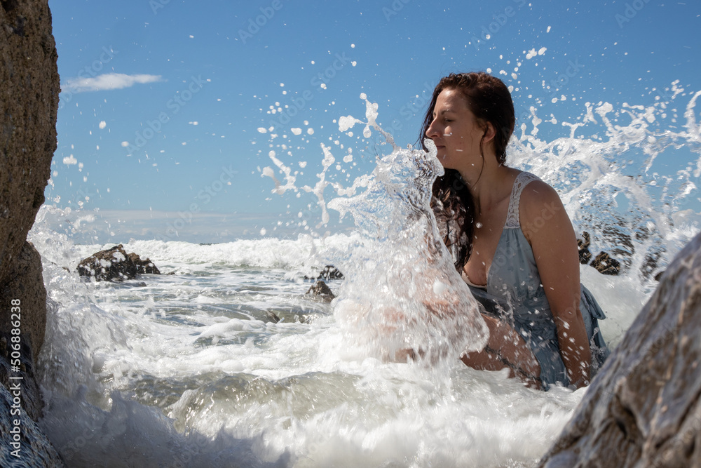 Obraz premium A woman in a blue dress at the beach on a sunny day. The blue sky has some cloud cover. The model sits amongst the rocks as the sea washes waves over her