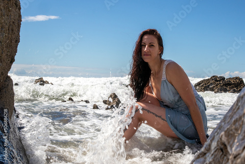 A woman in a blue dress at the beach on a sunny day. The blue sky has some cloud cover. The model sits amongst the rocks as the sea washes waves over her