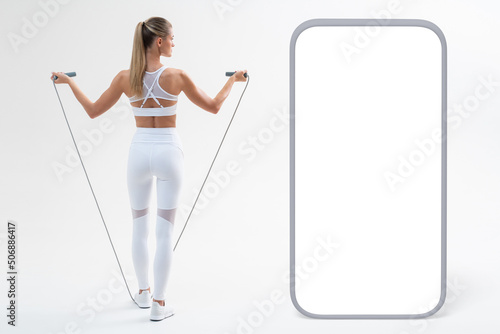 Back view of blonde woman with skipping rope doing cardio workout next to big phone mock up photo