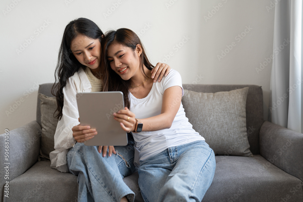 Happy young lesbian couple hugging having fun using digital tablet relaxing on couch at home. Two smiling asian women friends holding computer looking at screen enjoying surfing online watching videos Happy young lesbian couple hugging having fun using digital tablet relaxing on couch at home. Two smiling asian women friends holding computer looking at screen enjoying surfing online watching videos