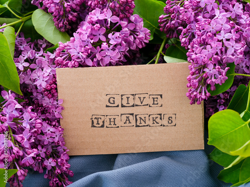 A piece of cardboard with words Give Thanks on it next to some lilac flowers and leaves