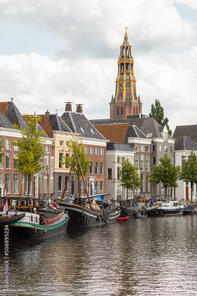 Historic ships, canal houses and warehouses on the old harbor at the Hoge der A with the tower of the historic Der Aa church in Groningen in the background.