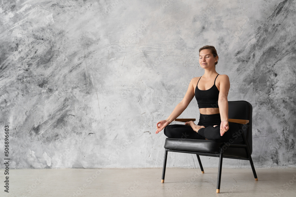 Young woman practicing yoga in lotus pose on black chair at home, meditation concept
