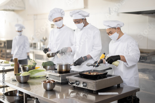 Multiracial team of cooks in uniform and face masks cooking meals for a restaurant in the kitchen. Concept of teamwork at restaurant during pandemic. Latin, Asian and European guys cooking together © rh2010