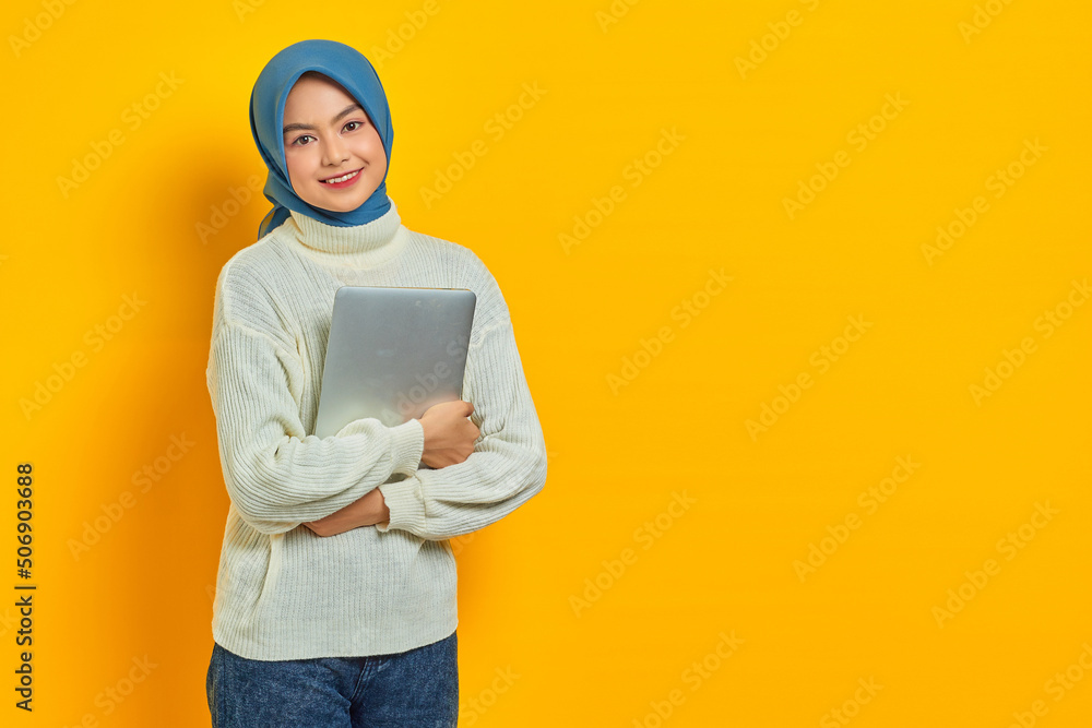 Portrait of smiling beautiful asian woman in white sweater and hijab holding a laptop and looking at camera isolated over yellow background. people religious lifestyle concept Portrait of smiling beautiful asian woman in white sweater and hijab holding a laptop and looking at camera isolated over yellow background. people religious lifestyle concept