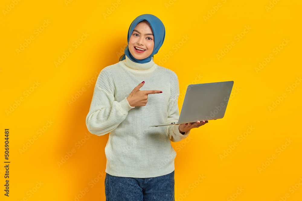 Cheerful beautiful asian woman in white sweater and hijab pointing fingers at a laptop, showing something online isolated over yellow background. people religious lifestyle concept Cheerful beautiful asian woman in white sweater and hijab pointing fingers at a laptop, showing something online isolated over yellow background. people religious lifestyle concept
