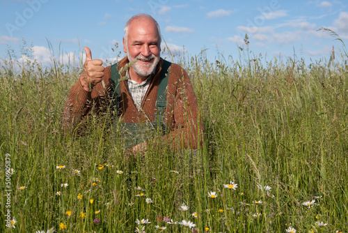 A friendly old farmer in his organically farmed meadow smiles at the camera and gives his thumbs up. Organic farming is better, it protects biodiversity, the soil and keeps the climate impact low. photo