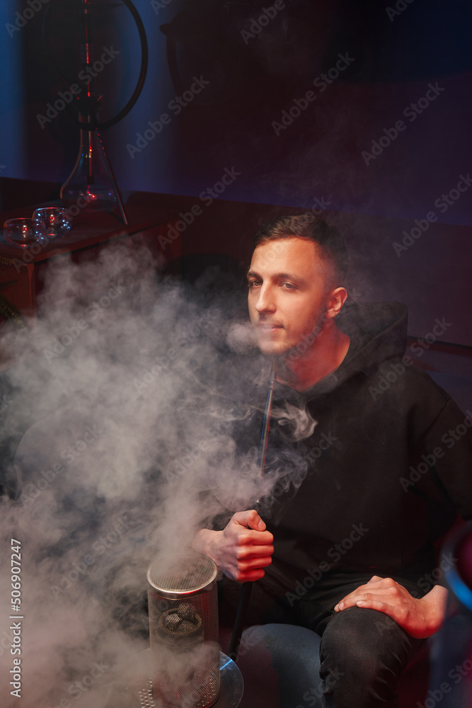 a young man smokes a hookah in a dark hookah, exhales a large cloud of white smoke.