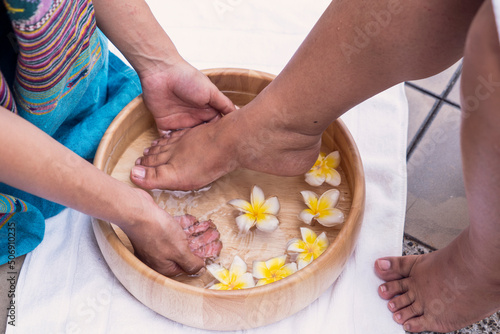 Foot spa ,wellness and Relax concept The masseur washes his bare feet in a wooden tub with frangipani flowers and salt scrub and soap near swimming pool at the resort and spa .