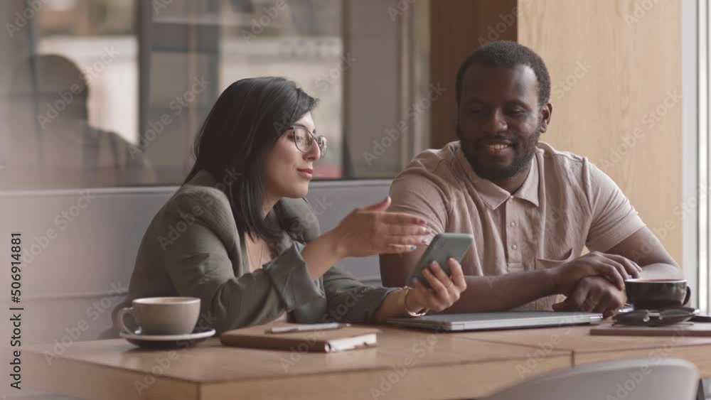 Medium slowmo of smiling young multiethnic business couple discussing ideas of corporate project while working together in cozy cafeteria near their office