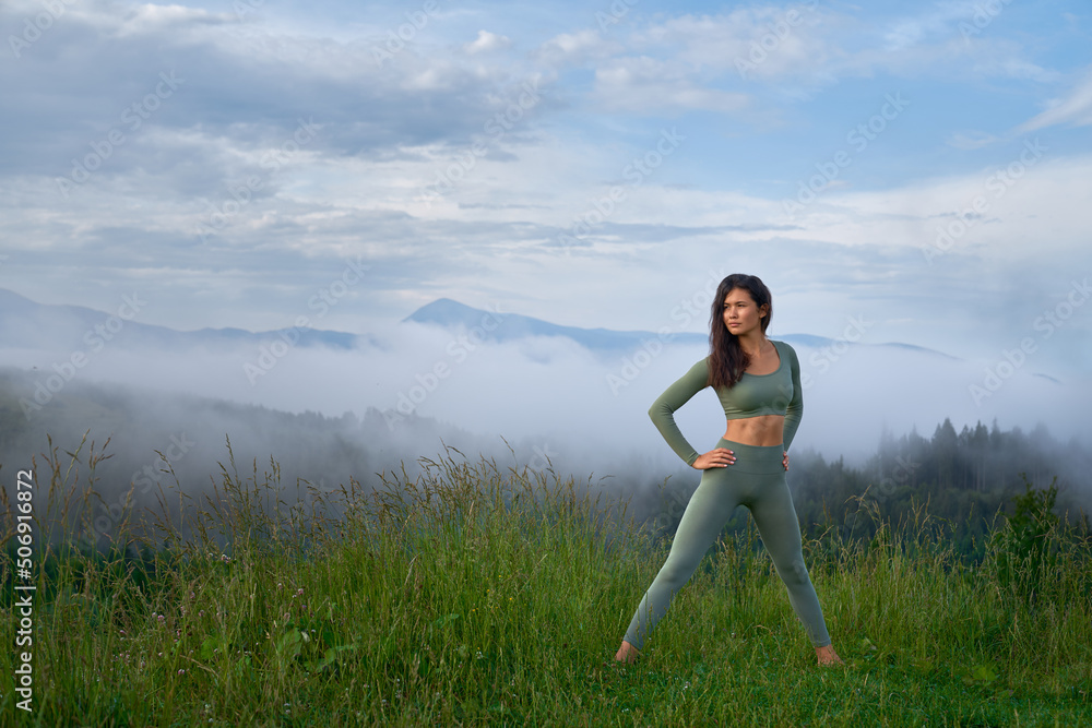 Beautiful woman looking at side, standing by feet go green grass, preparing for practice of yoga in morning. Pretty female with hands on hips, looking at side, stretching with mountains on background. Beautiful woman looking at side, standing by feet go green grass, preparing for practice of yoga in morning. Pretty female with hands on hips, looking at side, stretching with mountains on background.