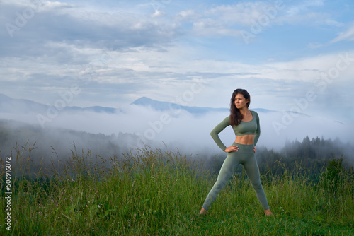 Beautiful woman looking at side, standing by feet go green grass, preparing for practice of yoga in morning. Pretty female with hands on hips, looking at side, stretching with mountains on background.