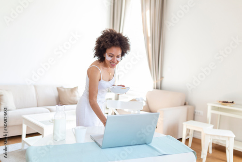 young amazing woman sitting indoors at the table with laptop holding corn flakes. Looking at laptop computer and talking to her friends via video call.