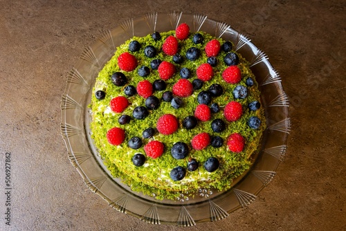 Top view of a round homemade fruit cake with red raspberries, blueberries and green top made of spinach placed on glass dish on brown surface.