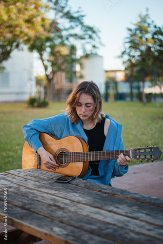 Girl practicing a song with her guitar while reading music sheet on her phone. She is sitting in a wooden picnic area.