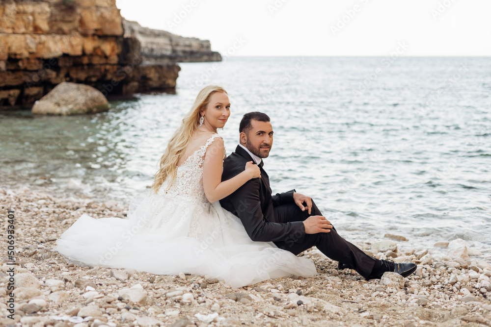 Portrait of young blonde bride in white dress and brunet groom in suit embracing and sitting on rocky sea coast in Italy side view, seascape background. Beautiful and romantic wedding, happy couple