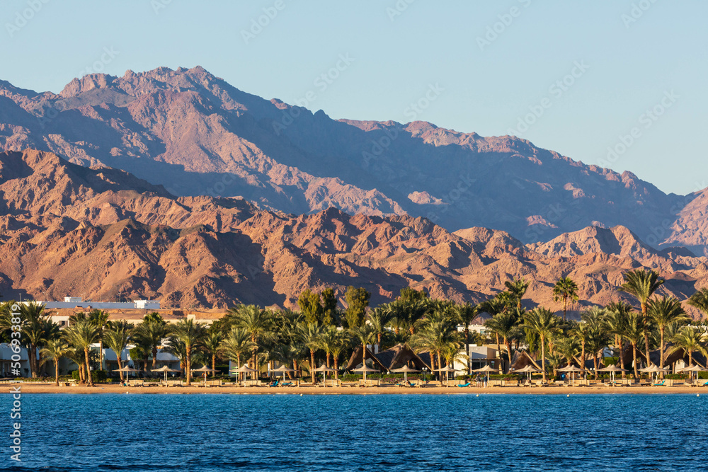 The coast of Lagoon beach with hotels, palm trees and mountains. View across the sea. Dahab, Egypt