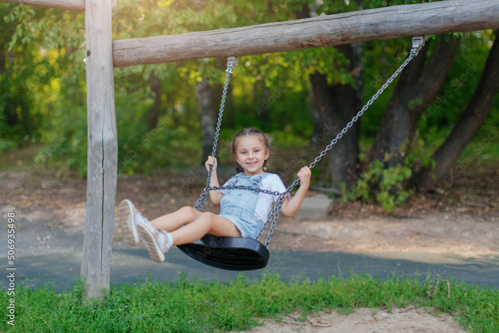A child plays on an outdoor playground on a summer day. An active happy little girl is riding on a swing. Healthy summer activity for children in warm weather. The little cute baby is rocking.