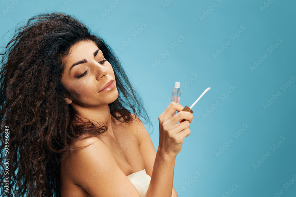 Attractive happy curly Latin lady looks at lip gloss tube posing isolated over pastel blue background. Cosmetic lips product ad Natural beauty Makeup concept Studio portrait Copy space Attractive happy curly Latin lady looks at lip gloss tube posing isolated over pastel blue background. Cosmetic lips product ad Natural beauty Makeup concept Studio portrait Copy space