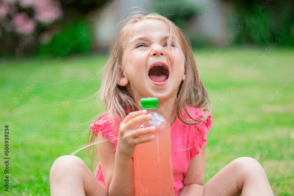 Drink with electrolytes in a transparent bottle in the hands of a happy child on a green lawn in a pink T-shirt Drink with electrolytes in a transparent bottle in the hands of a happy child on a green lawn in a pink T-shirt