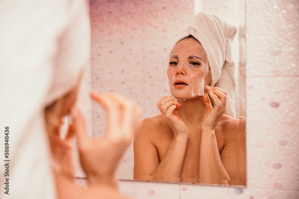 Young beautiful woman using a moisturizing facial mask after taking a bath. Pretty attractive girl in a towel on her head stands in front of a mirror in a home bathroom. Daily hygiene and skin care Young beautiful woman using a moisturizing facial mask after taking a bath. Pretty attractive girl in a towel on her head stands in front of a mirror in a home bathroom. Daily hygiene and skin care