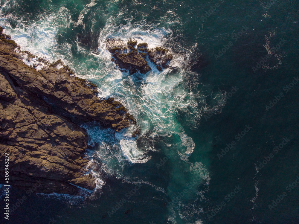 Aerial view of the ocean. The water is bright turquoise with white foamy waves crashing against the stone shore. Geology, geography. Travel, tourism, environmental protection.